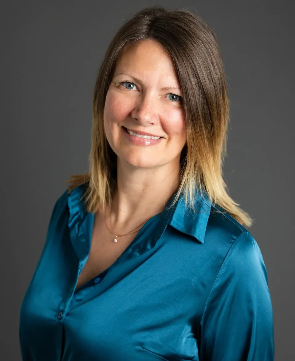 Smiling woman with long hair in a teal blouse against a gray background.